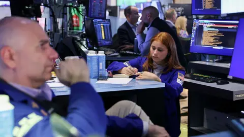 Getty Images In the left corner, a bald male trader, and a woman with auburn hair, also a trader, in the middle, holding a pen and writing into a notebook, on the floor of the New York Stock Exchange.